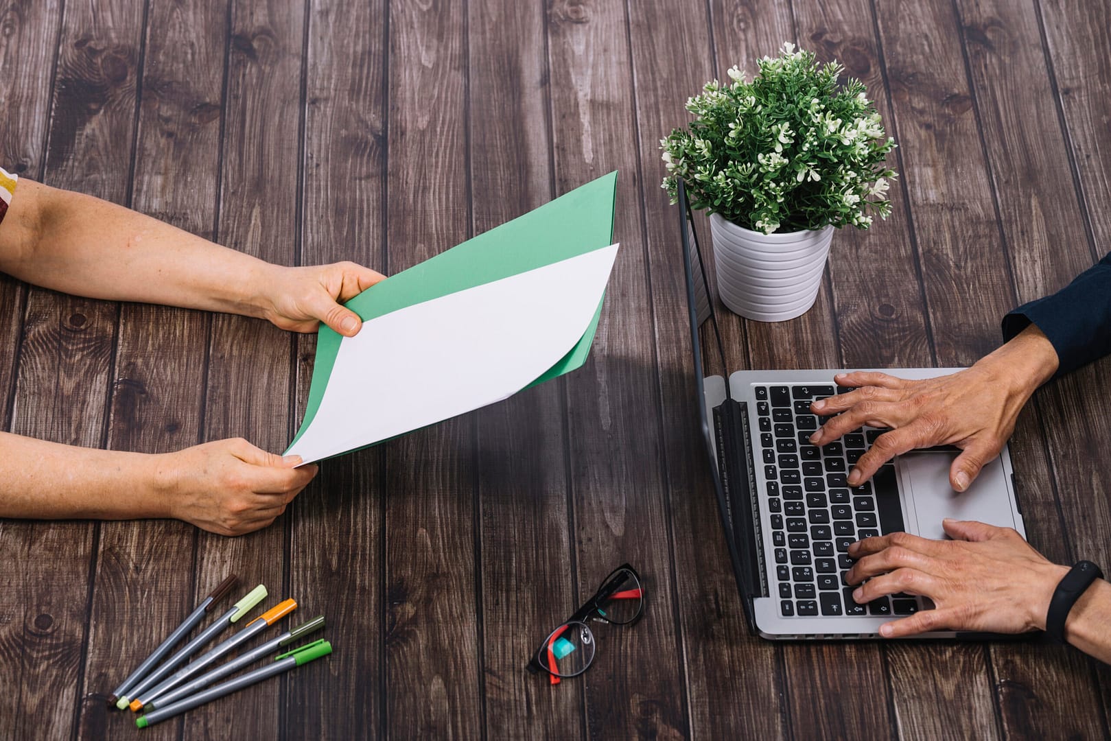 person typing laptop with his colleague holding blank white green blank paper wooden table 1 scaled
